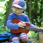 Ein Kind in bunter Kleidung spielt sitzend auf der Gitarre im Wald